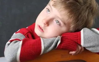 A young boy with light brown hair and blue eyes leans his head and arms on the back of a wooden chair, gazing upward thoughtfully. He wears a red and gray striped sweater—perhaps lost in thought about ADHD symptoms—while a blackboard sits behind him. - The Parker Group A young boy with light brown hair and blue eyes leans his head and arms on the back of a wooden chair, gazing upward thoughtfully. He wears a red and gray striped sweater—perhaps lost in thought about ADHD symptoms—while a blackboard sits behind him.