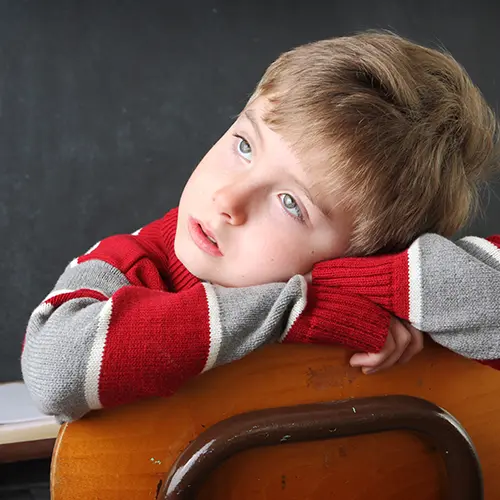 A young boy with light brown hair and blue eyes leans his head and arms on the back of a wooden chair, gazing upward thoughtfully. He wears a red and gray striped sweater—perhaps lost in thought about ADHD symptoms—while a blackboard sits behind him. A young boy with light brown hair and blue eyes leans his head and arms on the back of a wooden chair, gazing upward thoughtfully. He wears a red and gray striped sweater—perhaps lost in thought about ADHD symptoms—while a blackboard sits behind him.