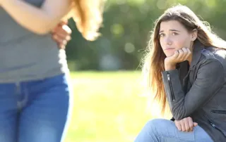 A young woman with long hair, wearing a gray jacket and jeans, sits on a bench outdoors, resting her chin on her hand and looking sad—perhaps struggling with ADHD symptoms. Another woman walks away in the foreground amid sunlight and green bushes. - The Parker Group A young woman with long hair, wearing a gray jacket and jeans, sits on a bench outdoors, resting her chin on her hand and looking sad—perhaps struggling with ADHD symptoms. Another woman walks away in the foreground amid sunlight and green bushes.