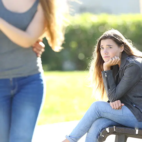 A young woman with long hair, wearing a gray jacket and jeans, sits on a bench outdoors, resting her chin on her hand and looking sad—perhaps struggling with ADHD symptoms. Another woman walks away in the foreground amid sunlight and green bushes. A young woman with long hair, wearing a gray jacket and jeans, sits on a bench outdoors, resting her chin on her hand and looking sad—perhaps struggling with ADHD symptoms. Another woman walks away in the foreground amid sunlight and green bushes.