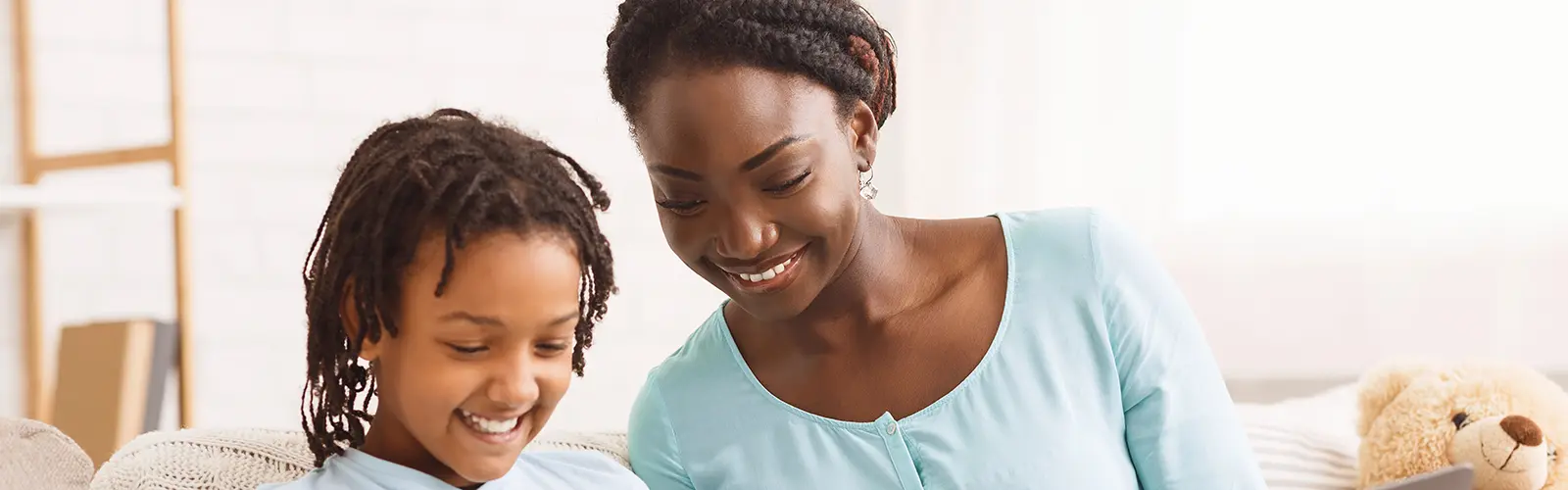 A smiling woman and a young girl with braided hair sit close together on a light-colored couch, enjoying time together. Both wear light blue tops. A teddy bear rests beside them, creating a softly lit, homely scene—ideal for relaxing after neurofeedback sessions.