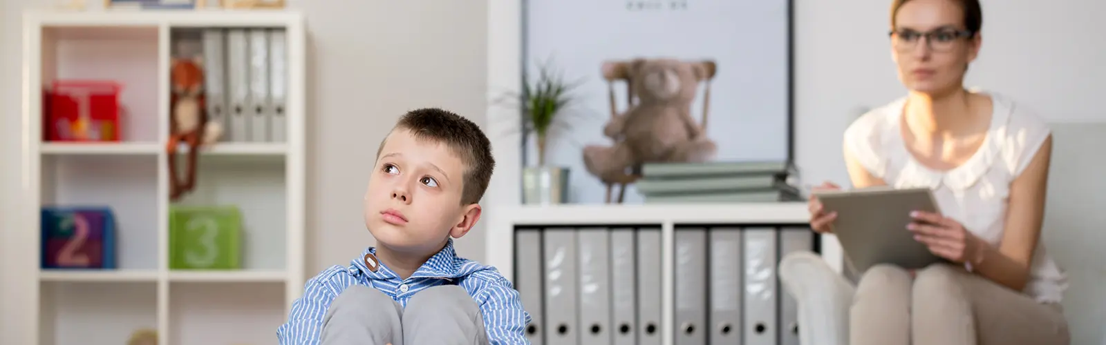 A young boy sits on the floor looking up thoughtfully, while a woman with glasses, possibly a neurofeedback therapist, sits behind him on a chair. The setting appears to be an office or therapy room with shelves, a plant, and a teddy bear in the background.