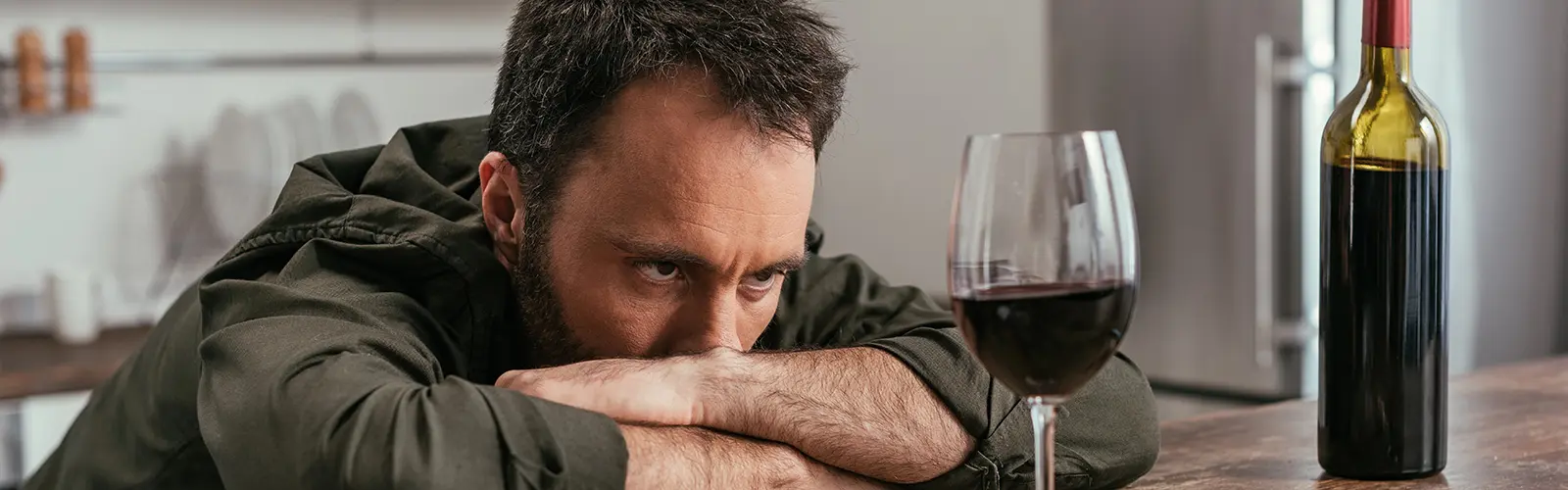 A man with a tired, troubled expression rests his arms and head on a table, staring at a glass of red wine. In the kitchen, the scene suggests sadness or contemplation—perhaps reflecting on neurofeedback and ways to find relief.