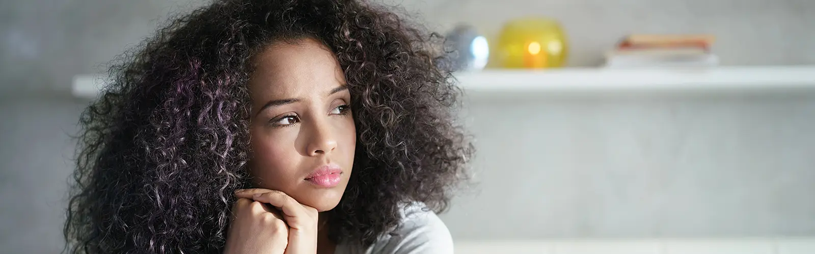 A young woman with curly dark hair rests her chin on her hands, gazing thoughtfully to the side. She appears pensive, perhaps reflecting on her recent neurofeedback session. The background is blurred, showing a white shelf with books and decorative objects.