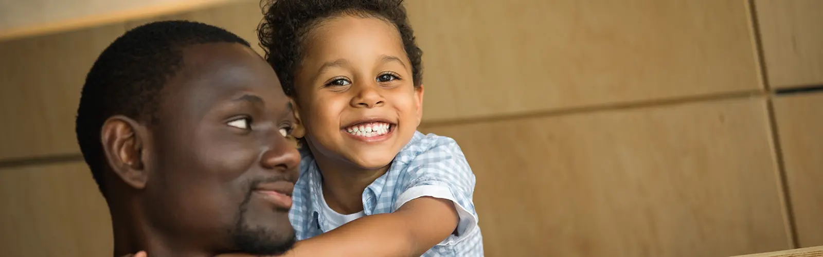 A young boy with curly hair and a big smile hugs an adult man from behind. The man, perhaps after a neurofeedback session, looks to the side, smiling softly. Indoors with a beige wall in the background, both appear happy and affectionate.