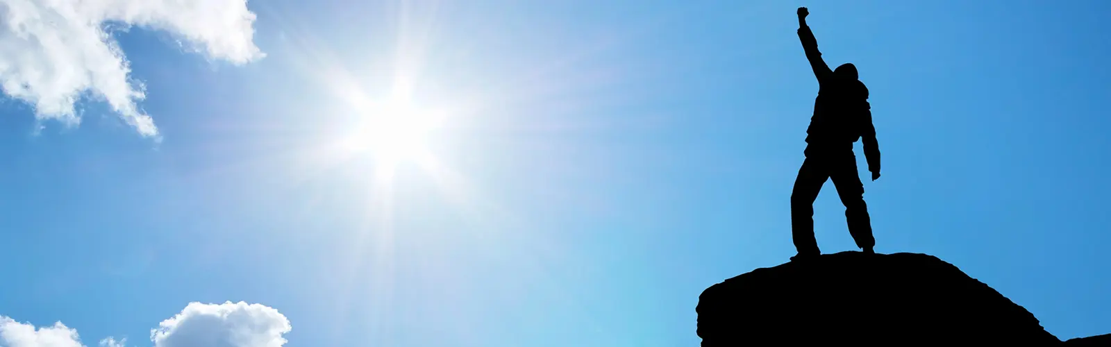 A silhouetted figure stands triumphantly on a mountaintop with one arm raised toward the sky. Like neurofeedback guiding the mind, the sun shines brightly in the blue sky, symbolizing achievement, success, and reaching new heights.