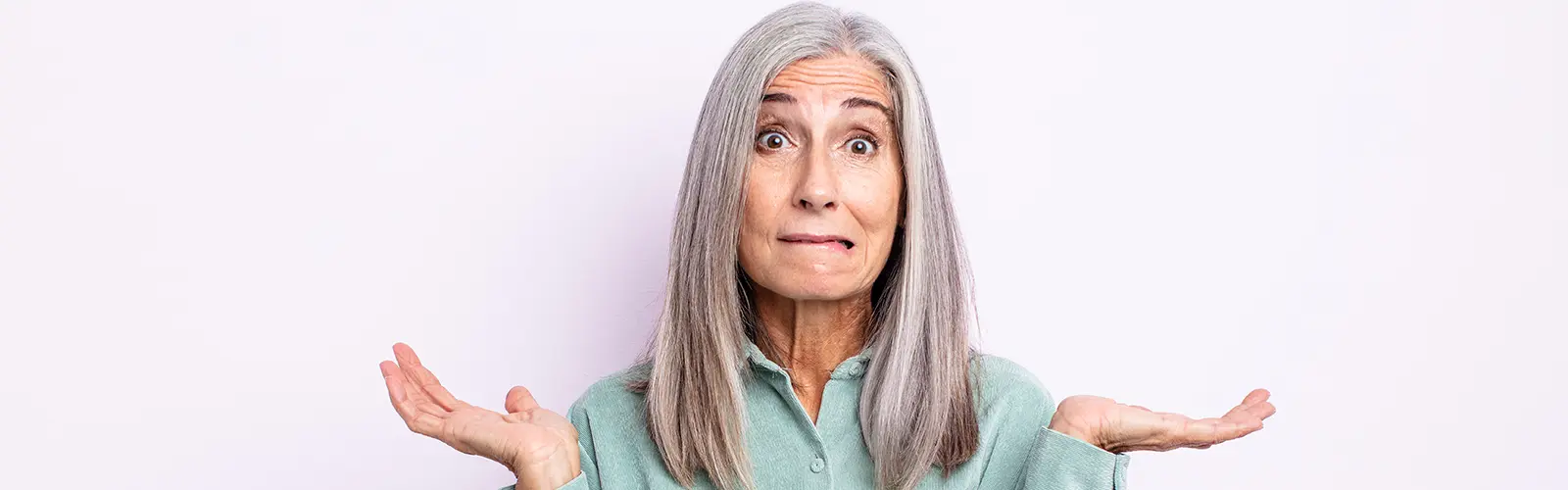 An older woman with long gray hair wearing a light green shirt stands against a light background, raising both hands with palms up in a confused expression, as if unsure about neurofeedback.