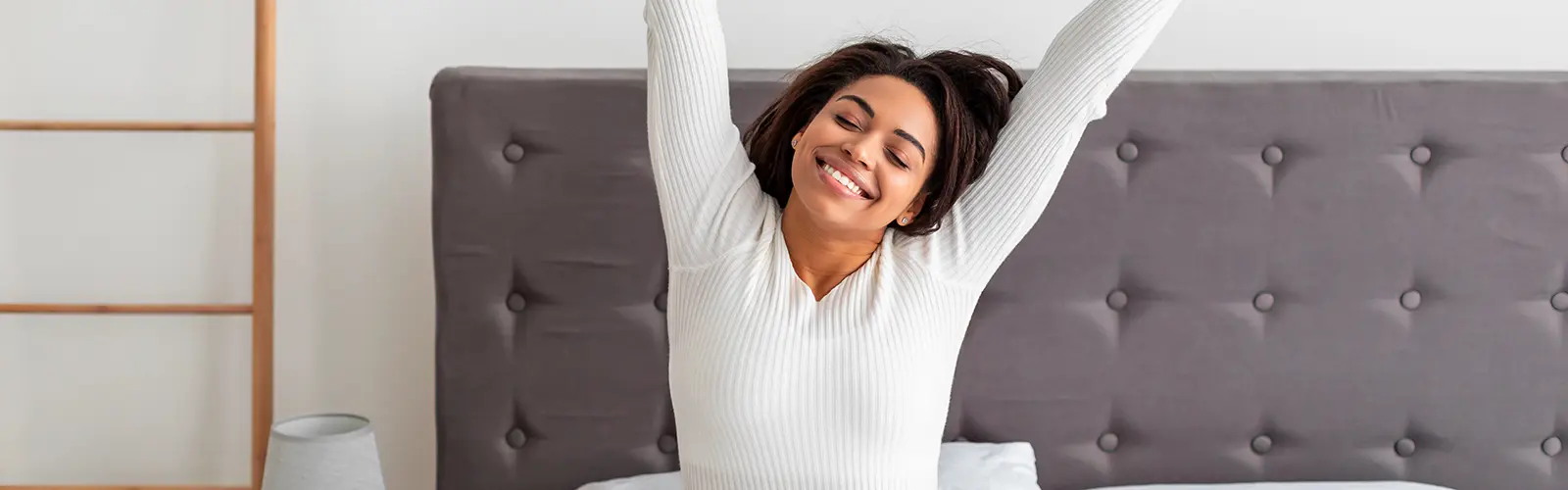 A woman with medium skin and dark hair, wearing a white long-sleeve top, sits on a bed and stretches her arms upward while smiling, enjoying a bright, minimal room—perhaps after a peaceful morning neurofeedback session.