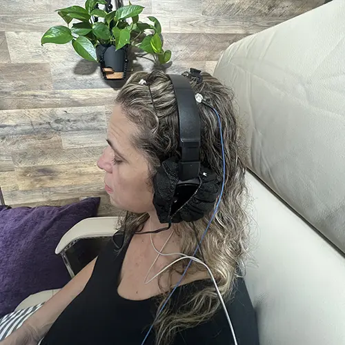 A woman with wavy hair sits on a beige chair, wearing a black headset with electrodes and wires for a Brain Map QEEG. She is in profile, facing left. A small green potted plant sits on a wooden wall in the background. A woman with wavy hair sits on a beige chair, wearing a black headset with electrodes and wires for a Brain Map QEEG. She is in profile, facing left. A small green potted plant sits on a wooden wall in the background.
