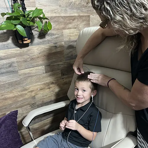 A young boy sits in a beige chair, smiling, while a woman attaches electrodes to his scalp for neurofeedback. The boy holds wires connected to the electrodes. A plant hangs on the wood-paneled wall in the calm, clinical setting. A young boy sits in a beige chair, smiling, while a woman attaches electrodes to his scalp for neurofeedback. The boy holds wires connected to the electrodes. A plant hangs on the wood-paneled wall in the calm, clinical setting.