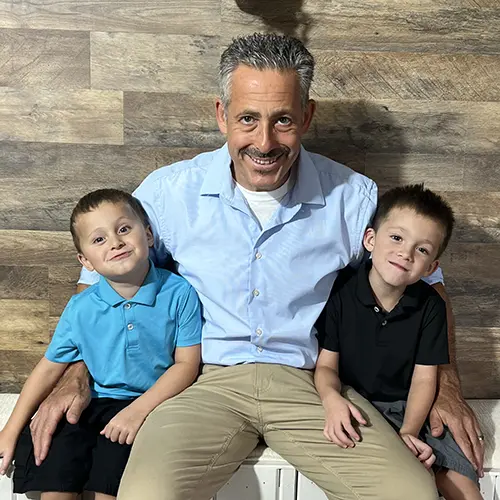 A smiling man with gray hair and a mustache sits on a bench, wearing a light blue shirt and tan pants. He has his arms around two young boys—one in a blue polo and shorts, the other in a black polo and shorts. Wood-patterned wall background. A smiling man with gray hair and a mustache sits on a bench, wearing a light blue shirt and tan pants. He has his arms around two young boys—one in a blue polo and shorts, the other in a black polo and shorts. Wood-patterned wall background.