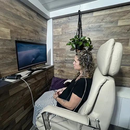 A woman sits in a reclining chair with medical cables attached to her arms, experiencing the Parker Group Method while watching an ocean scene on a monitor. She relaxes in a cozy room with wood-paneled walls and a hanging green plant above her. A woman sits in a reclining chair with medical cables attached to her arms, experiencing the Parker Group Method while watching an ocean scene on a monitor. She relaxes in a cozy room with wood-paneled walls and a hanging green plant above her.