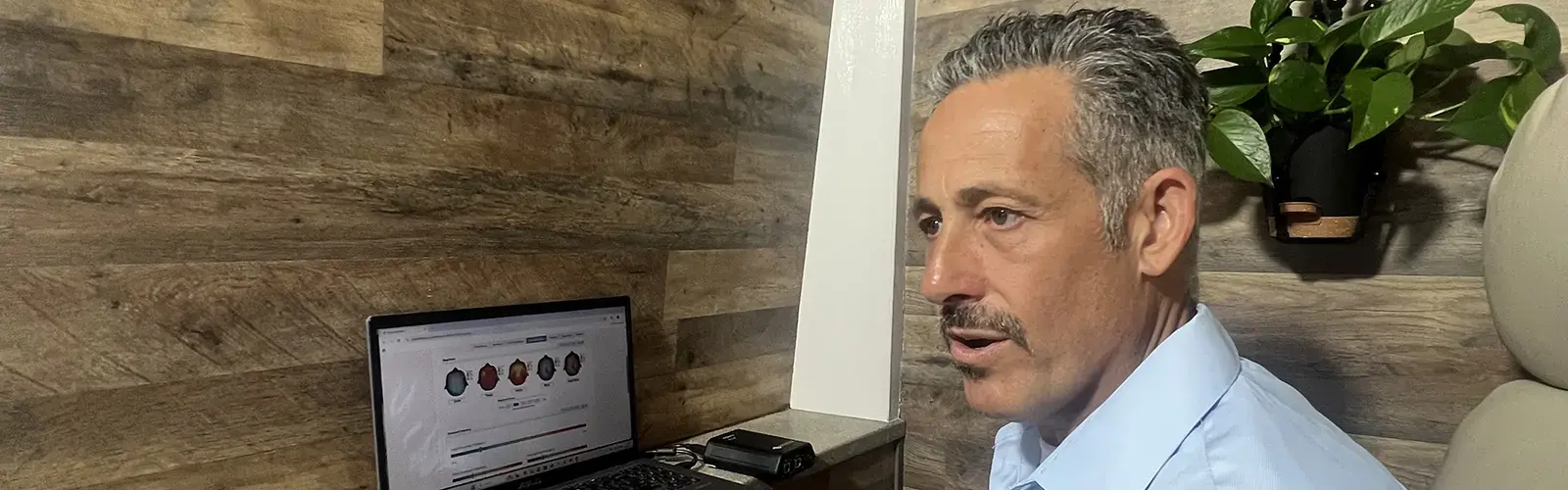 A man with short, curly gray hair and a mustache sits at a desk, looking at a laptop displaying charts and faces, possibly related to Neurofeedback or EEG biofeedback. He wears a light blue shirt in his wood-paneled home office with a green potted plant.